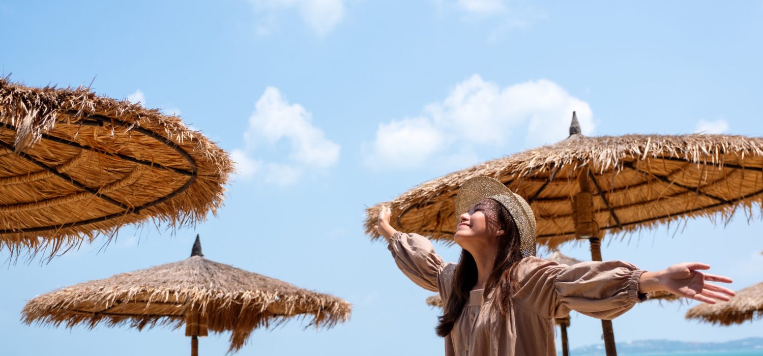 A beautiful young asian woman raising hands while strolling on the beach with the sea and blue sky background